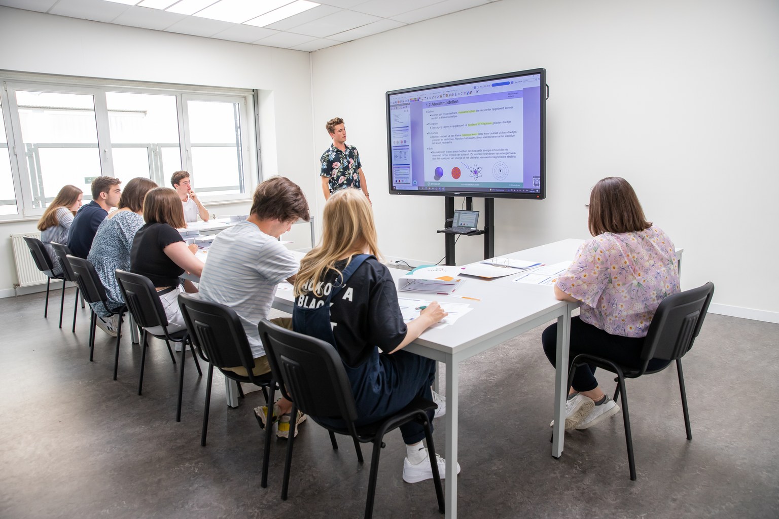 Een kleine groep studenten volgt les aan lange tafels, met de begeleider bij het scherm