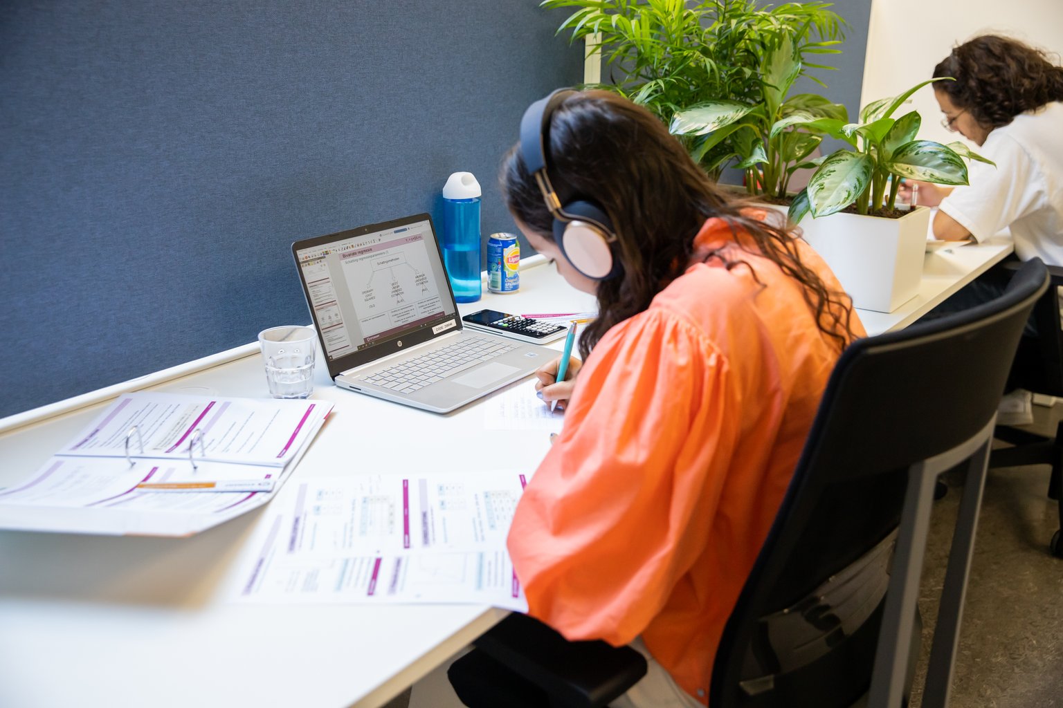 Een studente met koptelefoon volgt de online cursus op haar laptop en maakt notities